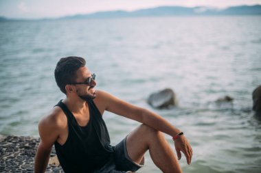Genç bir adam sahilde oturuyor. Gün batımında okyanus ufukta güneş gözlüğü yakışıklı adam bakar.