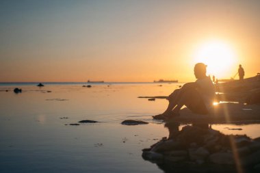 Genç bir adam bir taş denizin kenarında oturur. Adam düşünceli horizon adlı günbatımında görünüyor..