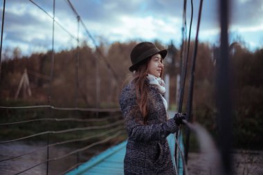 Genç kadın köyde büyük bir asma köprü üzerinde. Kız bir palto ve şapka bir sonbahar bulutlu günde bir ahşap köprü üzerinde yürüyor 