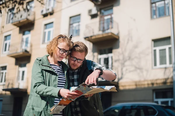 A young couple of tourists are looking for a road with a map in their hands in the city. The guy and the girl walk in a large European city with a map in their hands. Young tourists on the background of the city on a sunny spring day
