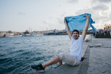 Rıhtımda denizin yanında genç bir adam sevinir ve güler.