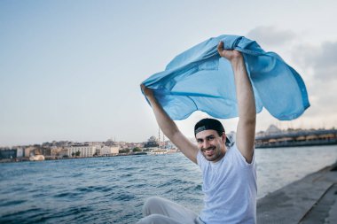 Rıhtımda denizin yanında genç bir adam sevinir ve güler.