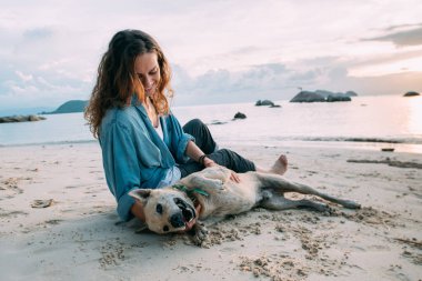 Kız gün batımında sahilde köpekle oynarken eğleniyor. Genç Avrupalı bir kadın deniz ve batan güneşin arka planında evcil bir köpekle yürüyor..