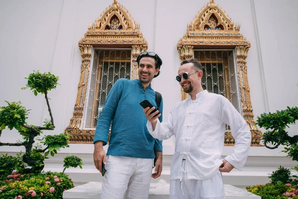 Men in a Buddhist temple read tourist information, look at a map on the phone. Young guys in simple national shirts against the background of the sights of the palace are watching an online guide on a smartphone. Tourists admire the architecture of S