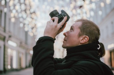 Elinde kamera olan bir adam kışın eski şehrin caddesini fotoğraflıyor. Arka planda sanatsal dekore edilmiş sokak lambalarının altında kamera ve eşarplı bir fotoğrafçı.
