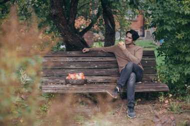 Erkek bir çiftçi bahçede bir bankta oturuyor. Ellerinde elma olan genç bir adam bir elma ağacının dallarının altındaki tahta bir bankta dinleniyor..