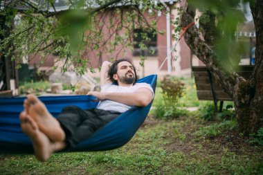 Bir adam bir kır evindeki hamakta dinleniyor. Genç bir adam hamakta yatıyor, yazın açık havada uyuyor kırsaldaki ağaçların yeşilliğinde.