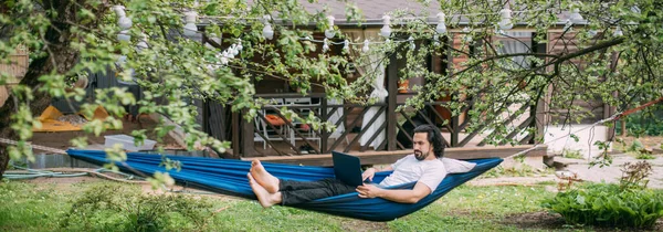 A man works with a laptop in a hammock in a country house. A young guy lies in a hammock with a computer, communicates online, learns remotely under green trees in the country in a summer