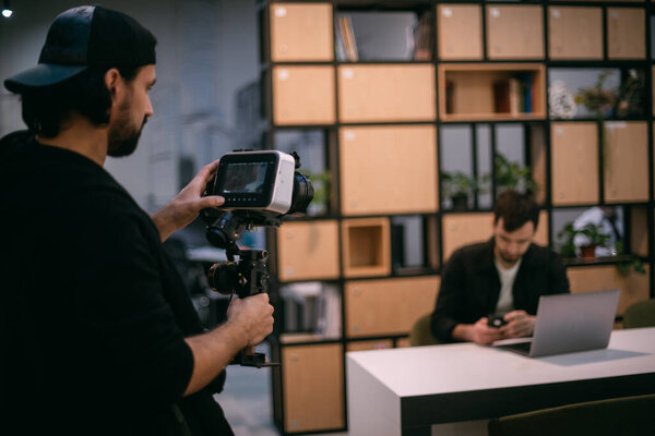 Filming a blog, workshop or live broadcast. A man with a camera shoots a guy at a laptop in a workspace. Professional cameraman and speaker make video in coworking