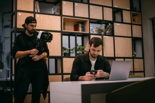 Filming a blog, workshop or live broadcast. A man with a camera shoots a guy at a laptop in a workspace. Professional cameraman and speaker make video in coworking