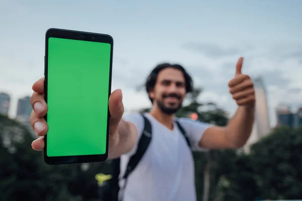 A man holds a phone with a green screen and stands against the backdrop of a metropolis. Young guy tourist with smartphone with chromakey on display. Phone in the foreground