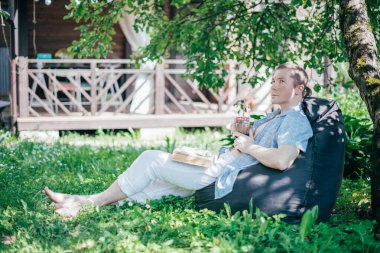 Bahçede bir adam dinleniyor ve bir sedirin üzerinde kitap okuyor. İzin günü ve dinlenme. Genç bir adam, güneşli bir günde elinde kitapla bir kır evinin gölgesinde bir ağacın altında yatıyor.