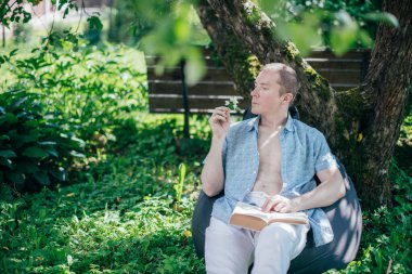 Bahçede bir adam dinleniyor ve bir sedirin üzerinde kitap okuyor. İzin günü ve dinlenme. Genç bir adam, güneşli bir günde elinde kitapla bir kır evinin gölgesinde bir ağacın altında yatıyor.