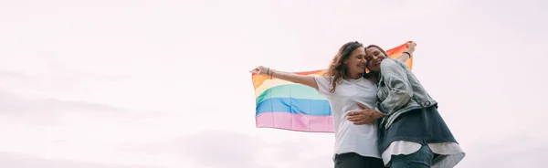 空に向かってビーチに虹の旗を持つ2人の女性 カップル若いレズビアンの女の子が虹のフラグを保持し 喜んで抱擁 レズビアンの女性のペア — ストック写真