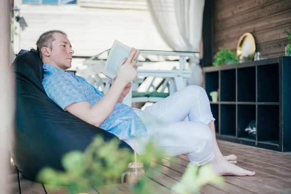 A man reads a book on the veranda. Young handsome guy lies on a ottoman with a book on the terrace of a country house on a sunny day