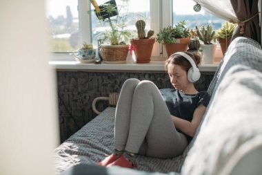 Elinde telefonla kanepede oturan bir kız. Genç bir kadın evde dinleniyor, kulaklıkla müzik dinliyor, akıllı telefondan video izliyor.