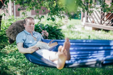 Bir adam hamakta dinleniyor ve kitap okuyor. İzin günü ve dinlenme. Genç yakışıklı bir adam, güneşli bir günde elinde kitapla bir hamakta, ağaçların gölgesinde yatıyor.