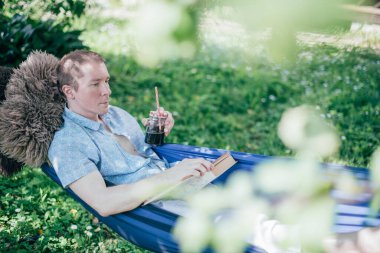 Bir adam hamakta dinleniyor ve kitap okuyor. İzin günü ve dinlenme. Genç yakışıklı bir adam, güneşli bir günde elinde kitapla bir hamakta, ağaçların gölgesinde yatıyor.
