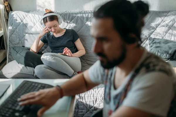 A man works at a table and a girl is resting on a sofa with a telephone. A young woman listens to music on headphones, watches a video. The guy at the computer at home