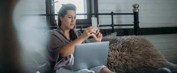 Woman works with laptop and phone on the bed in the bedroom. A young girl lies on a large bed at home, works online, speaks on the phone, communicates with clients