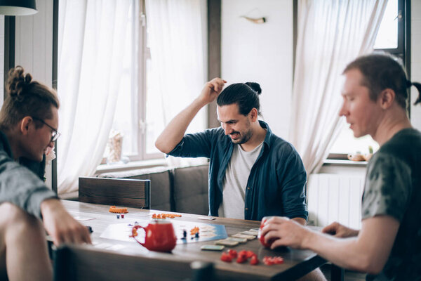 Friends play a board game in the living room. The company of young guys sits at a table and emotionally and cheerfully plays a card game on the weekend
