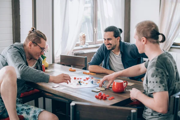 Friends play a board game in the living room. The company of young guys sits at a table and emotionally and cheerfully plays a card game on the weekend