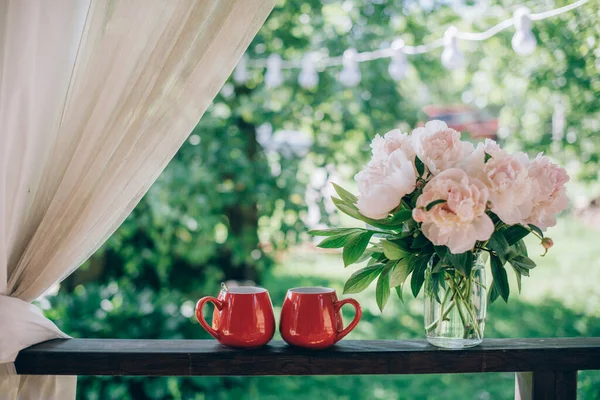 Still life of tea accessories and flowers. A pair of bright mugs of tea, a bouquet of peonies on the veranda on a background of a green garden on a bright day