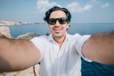 Bir adam deniz manzaralı kayalıklarda selfie çeker. Beyaz elbiseli yakışıklı bir adam kayalık bir sahilde okyanusa bakarken fotoğraflandı. # Denizin ötesindeki genç adam #