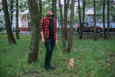 Bir adam kedisiyle parkta koşum takımı ile yürür. Evin yakınındaki yeşil parkta yürüyen genç bir adam. Kırmızı meraklı bir kediyle.