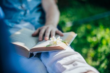 Bir adam hamakta dinleniyor ve kitap okuyor. Yakın plan. İzin günü ve dinlenme. Genç bir adam, güneşli bir günde, elinde bir kitapla, bir kır evinin gölgesinde, hamakta yatıyor.