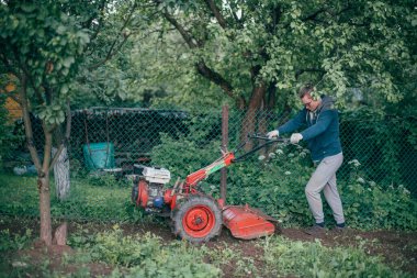 A man works in the garden, plows the soil with a cultivator. Young guy with a hand tractor digs up land for a vegetable garden in a country house clipart