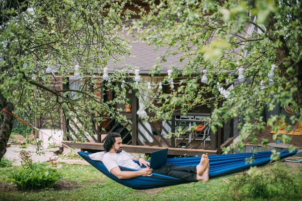 A man works with a laptop in a hammock in a country house. A young guy lies in a hammock with a computer, communicates online, learns remotely under green trees in the country in a summer