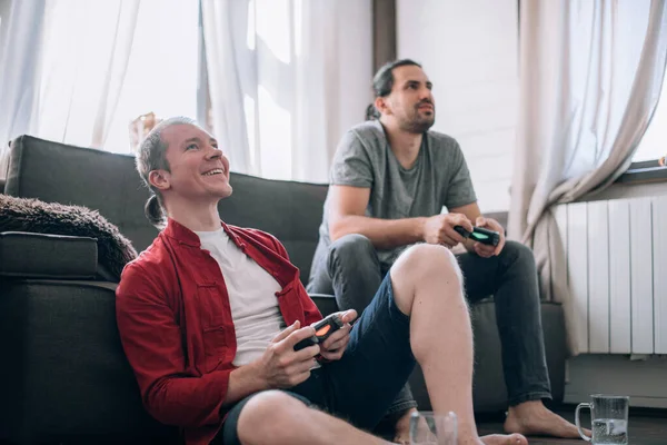 The guys are playing video games on the couch. Two young men with joysticks in their hands are enthusiastically playing a game on a console