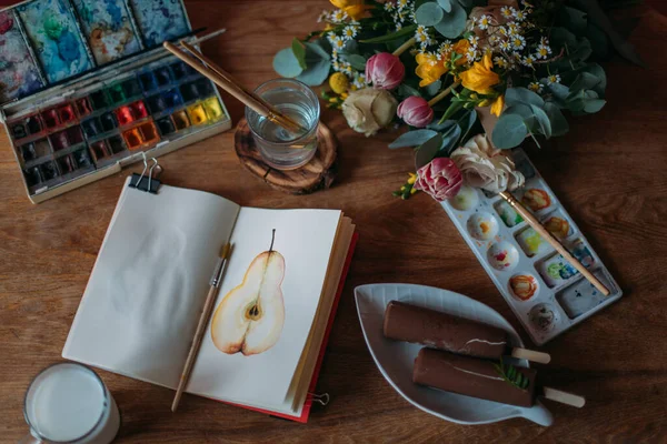 Artist\'s desktop with watercolors, palette, sketchbook. Still life. Wooden table with spring flowers bouquet, painting supplies, milk mug and ice cream