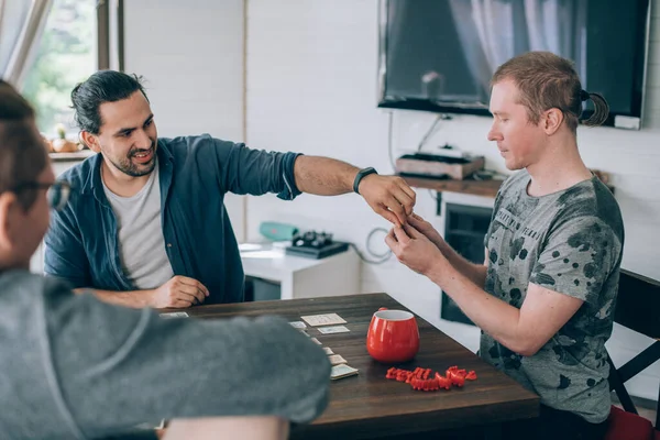 Friends play a board game in the living room. The company of young guys sits at a table and emotionally and cheerfully plays a card game on the weekend