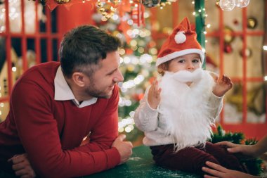 Baba ve küçük sevimli çocuk Noel Baba 'nın sahte sakallı Noel ağacının yanında Noel süslemeleri yapıyorlar. Komik bir çocuk beyaz bir sakal dener arka planda çelenkler ve festival ışıklarına karşı.