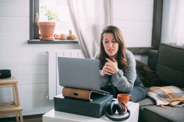 Bir kadın oturma odasında dizüstü bilgisayarla oturuyor, ekrana bakıyor, notlar alıyor, internetten yabancı bir dil öğreniyor, online eğitim kursları okuyor, internetten eğitim alıyor, bir müşteriye danışmanlık yapıyor.