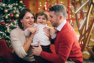 Noel süslemelerinde yeni yıl ağacının yanında çocuklu bir aile. Bebek, anne ve baba eğleniyor ve çelenk ve şenlik ışıklarına karşı oynuyorlar..