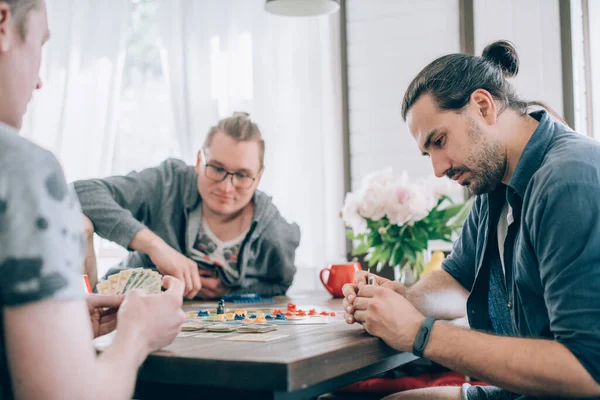 Friends play a board game in the living room. The company of young guys sits at a table and emotionally and cheerfully plays a card game on the weekend