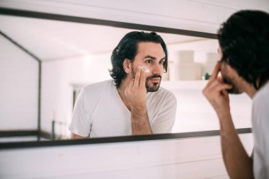 Yakışıklı adam yüz kremi sürüyor. Erkeklerin kişisel bakımı. Genç bir adam aynada kendine bakar ve yüzüne krema sürer.