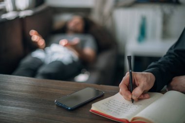 Psikoterapist ve hasta ofiste bir randevuda. Doktor 'un elleri not defterini yakın çekimde. Doktor müşterinin geçmişini kaydediyor. Bir erkek hasta, büyük, parlak bir ofiste, bir kanepede yatıyor.