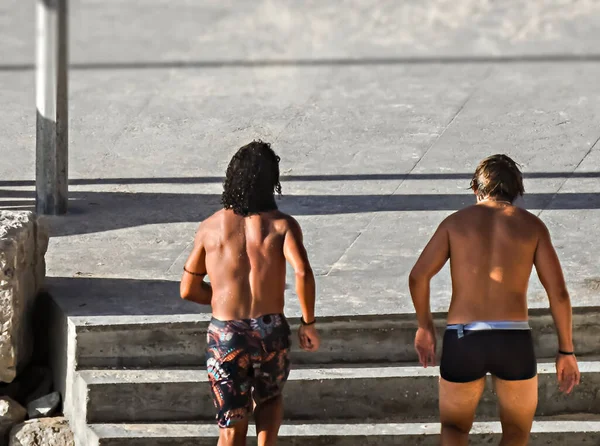 Two men in swimsuits running on a sidewalk with stairs near the beach