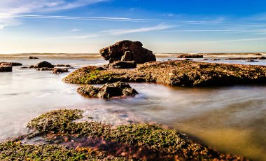 Kayalıklı deniz manzarası, durgun su ve birkaç beyaz bulutlu mavi bir gökyüzü.