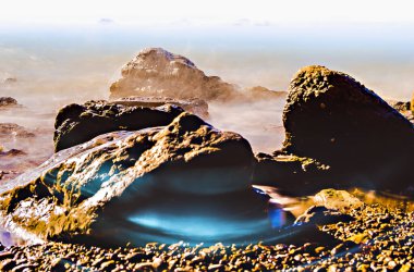 Kayalıklı deniz manzarası, durgun su ve birkaç beyaz bulutlu mavi bir gökyüzü.