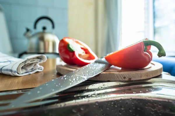 Grande faca de cozinha com gotas de água encontra-se entre dois pedaços de pimenta cortada, em uma placa de corte de madeira . — Fotografia de Stock