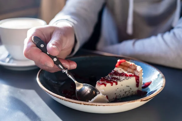 Νόστιμο cheesecake σπάει με ένα κουτάλι από ένα μεγάλο κομμάτι σε ένα όμορφο πιάτο, σε ένα θολό φόντο ένα φλιτζάνι καπουτσίνο. — Φωτογραφία Αρχείου