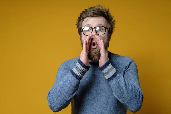 Man met een shaggy kapsel en een baard met handen op de lippen fluistert roddels en geruchten. Kopieerruimte. — Stockfoto