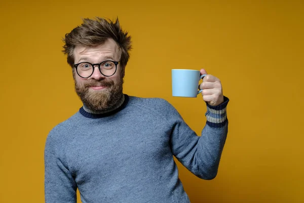 Hombre atractivo con un peinado peludo, gafas y un suéter viejo con una taza de bebida caliente tiene un buen estado de ánimo por la mañana temprano . — Foto de Stock