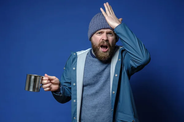 Upset man in camping clothes with a metal mug in hands, remembered that he had forgotten to take something important on the travel.