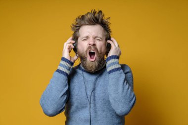 Eski bir süveter giymiş tatmin olmuş hipster kulaklıkla müzik dinliyor ve sarı bir arka planda yüksek sesle şarkı söylüyor..
